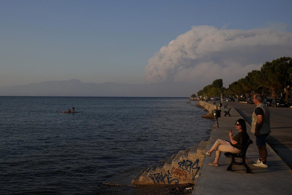 A man and woman enjoy the sea as a huge cloud of smoke, is seen in the background, fills the horizons at sun set over the island of Evia, where catastrophic wildfires continue to burn, in Arkitsa about 160 kilometers (100 miles) north of Athens, Saturday, Aug. 7, 2021. Wildfires rampaged through massive swathes of Greece's last remaining forests for yet another day Saturday, encroaching on inhabited areas and burning scores of homes, businesses and farmland. (AP Photo/Petros Karadjias)