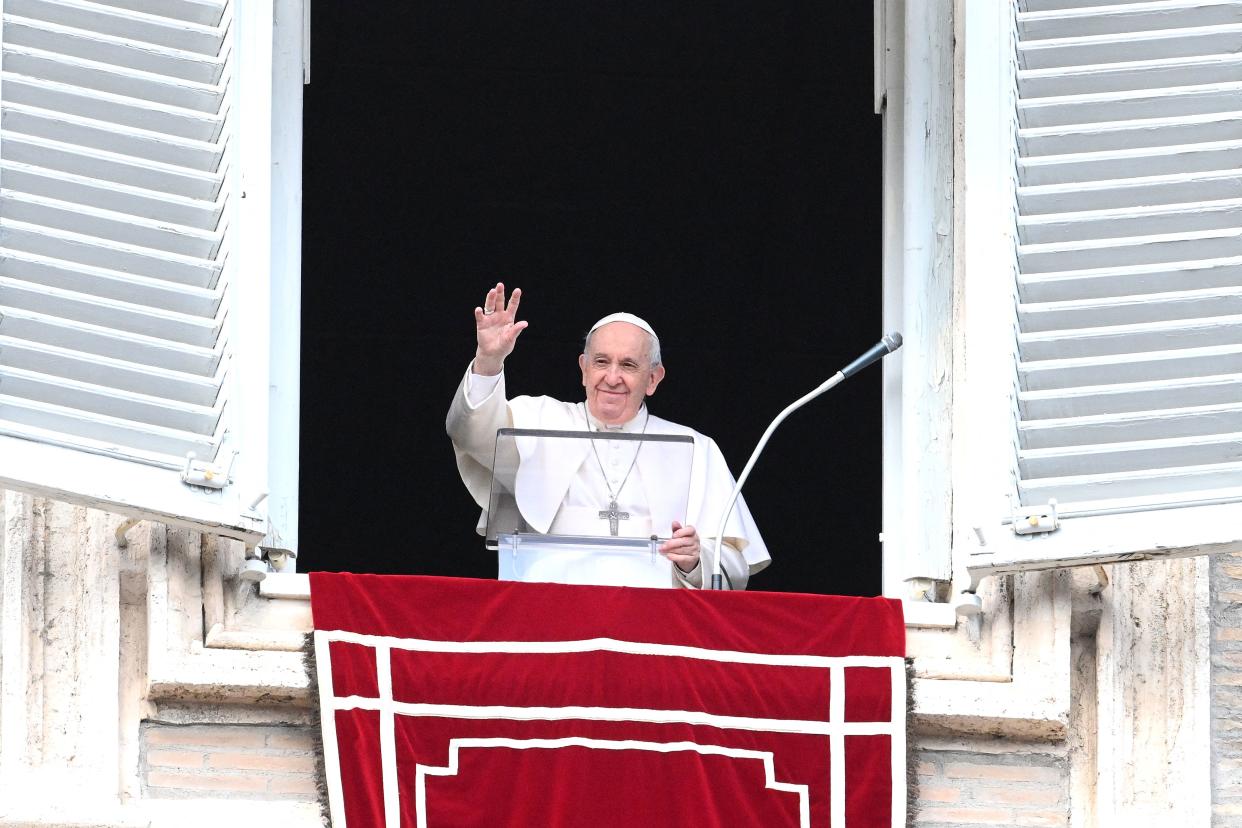 Pope Francis waves his hand from a large window with shutters thrown open.