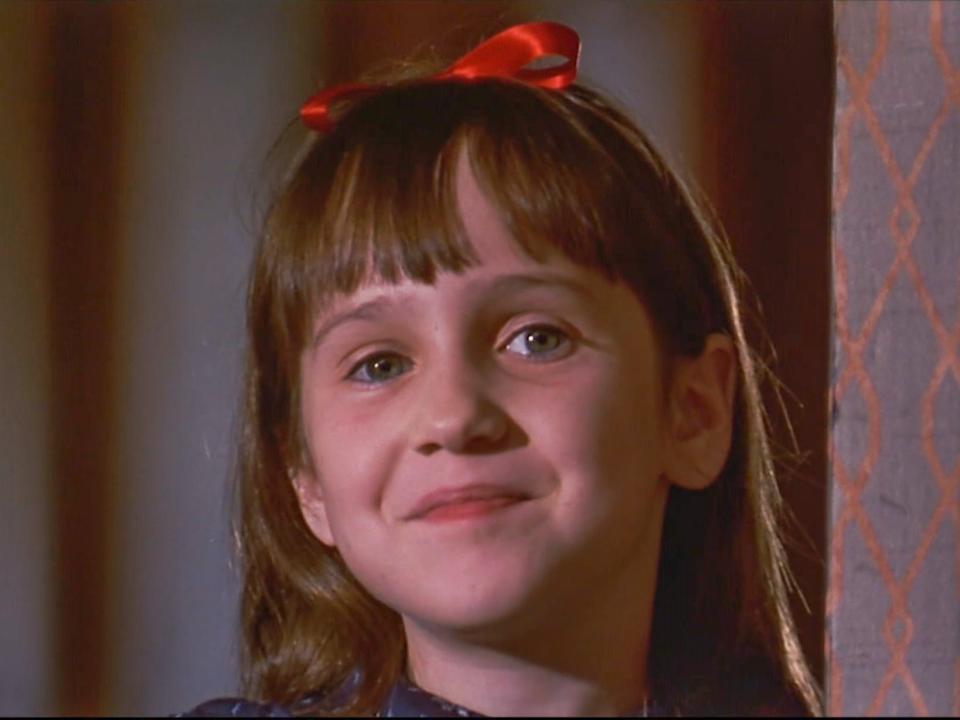 Mara Wilson as Matilda wearing a red bow in her hair and standing in a doorway