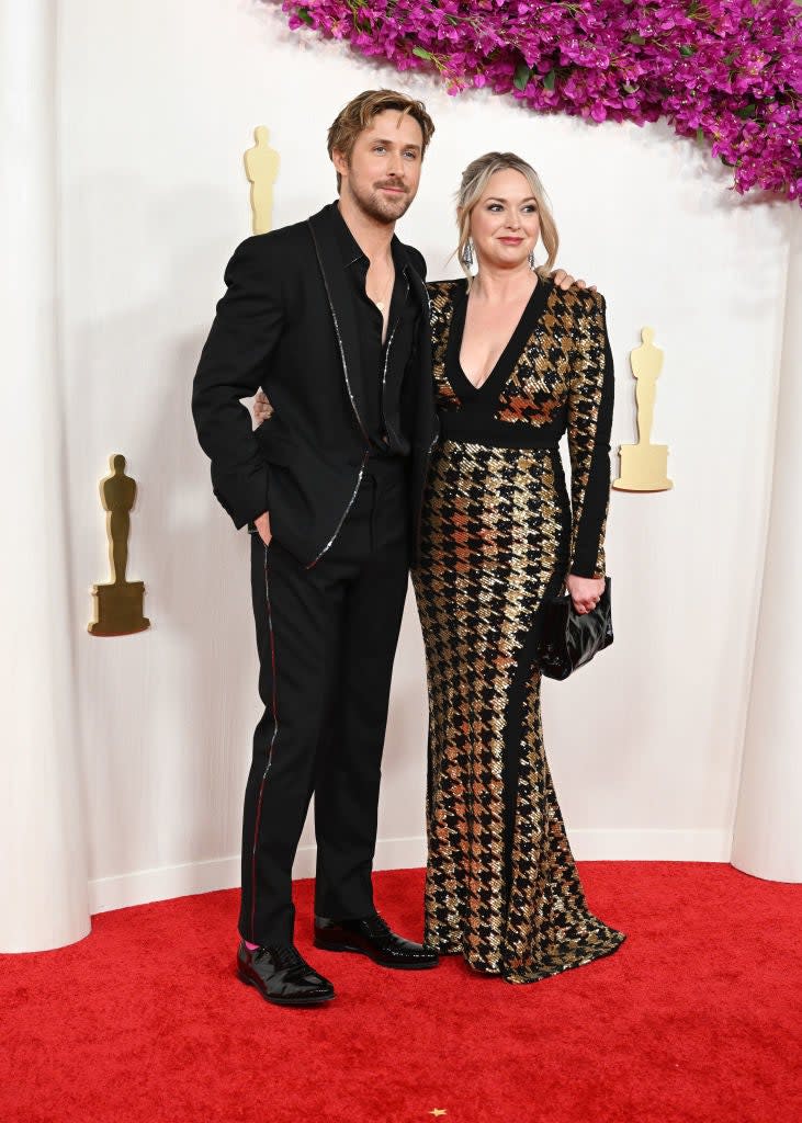 Two celebrities on red carpet; man in black suit with no tie, woman in gold patterned gown with clutch