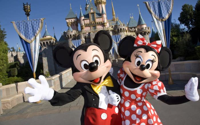 Mickey and Minnie Mouse waving to the camera in front of DIsneyland's princess castle.