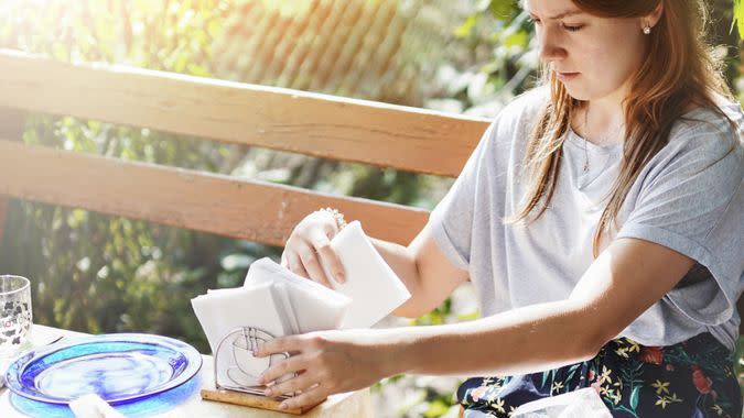 On a picnic terrace lit by the sun, a young girl with sunglasses on her head is laying out reusable glassware and paper napkins on a table.