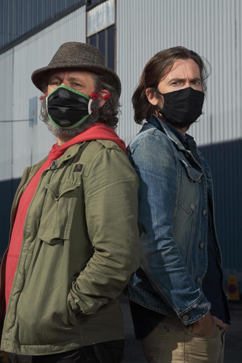 The actors Michael Sheen and David Tennant standing back to back in masks