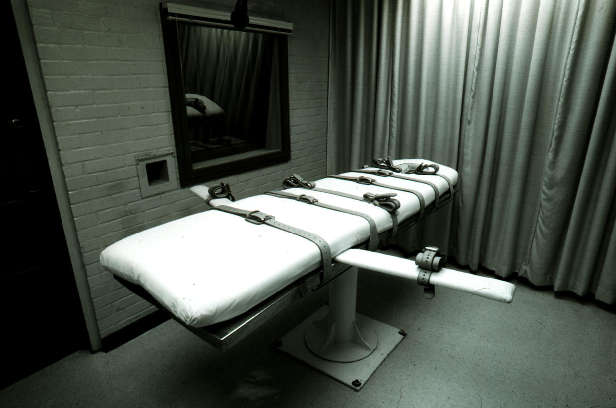 Monotone photo of an empty execution bed with several straps in a room with a brick wall next to a floor-to-ceiling curtain.