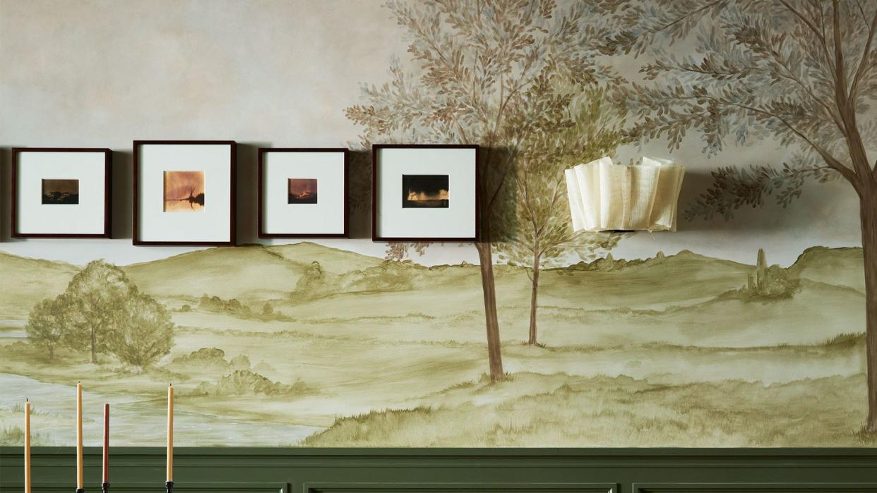 the bottom of a dining room wall is dark olive and the top is a green toned mural of a rural scene, a ruffled wall sconce, small framed artworks, travertine table with pedestal legs, wood chairs with off white fabric seats