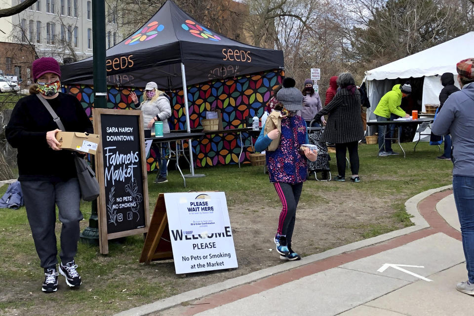 In this May 9, 2020 photo, customers pick up containers of vegetables, baked goods and other items at the downtown farmers' market in Traverse City, Mich. In normal times, the market would be crowded with shoppers browsing leisurely among dozens of stalls. But because of the coronavirus pandemic, they are asked to wear masks and observe social distancing while lining up for pre-ordered bags of food. (AP Photo/John Flesher)