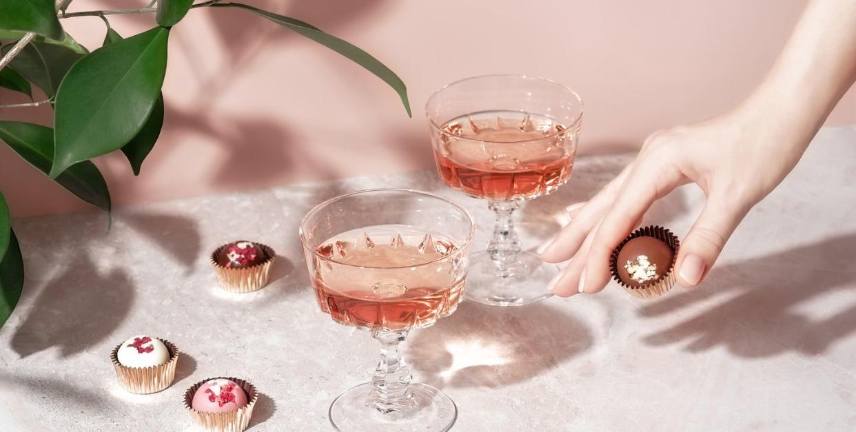 valentine's day party ideas, female hand holding chocolate candy over pastel pink marble table with crystal glasses of wine