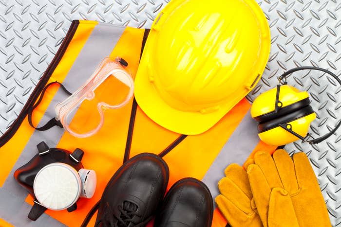 Safety gear on a metal surface, including a hard hat, safety vest, gloves, respirator mask, safety goggles, ear protection, and boots