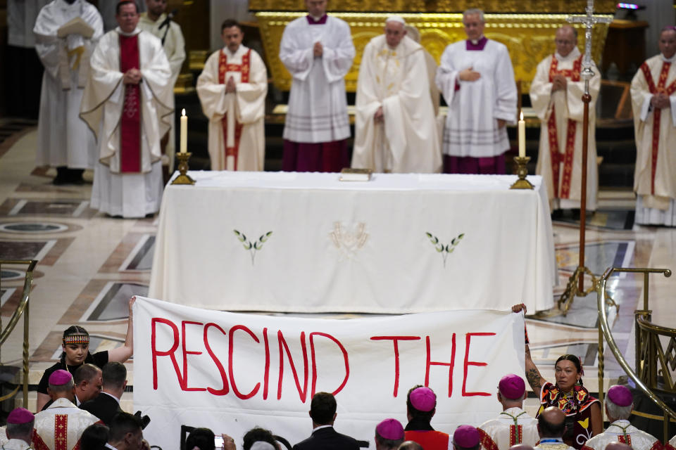 Protesters hold up a banner during a mass with Pope Francis, center top, at the National Shrine of Saint Anne de Beaupre, Thursday, July 28, 2022, in Saint Anne de Beaupre, Quebec. Pope Francis is on a "penitential" six-day visit to Canada to beg forgiveness from survivors of the country's residential schools, where Catholic missionaries contributed to the "cultural genocide" of generations of Indigenous children by trying to stamp out their languages, cultures and traditions. (AP Photo/John Locher)