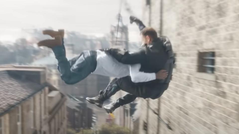 A man in a white shirt and blue jeans tackles a man in all black riding a zipline, near a bulilding