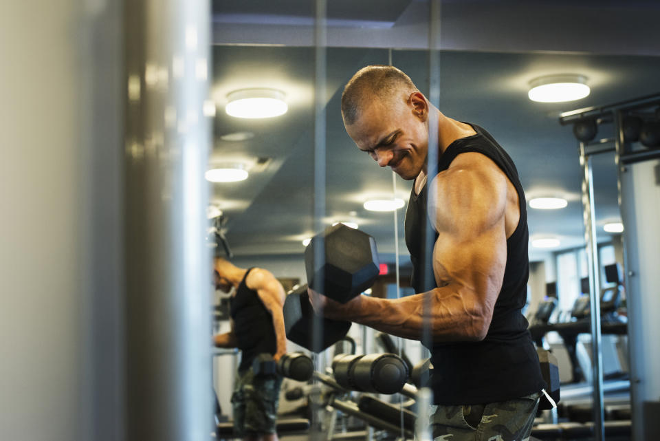 A person is lifting a dumbbell while looking in the mirror at a gym. They are wearing a sleeveless shirt and camo pants