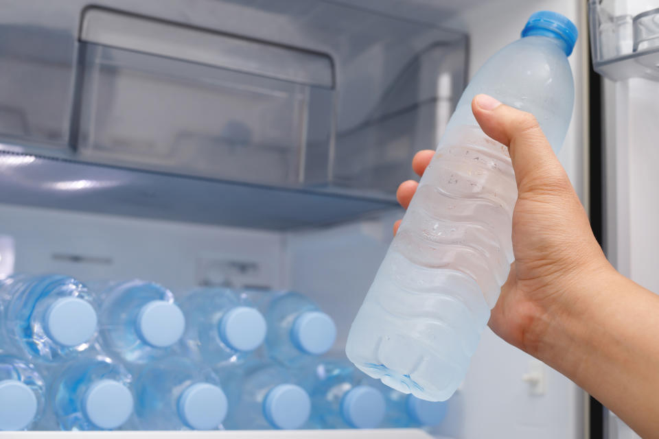 Hand holding a plastic water bottle, with more bottles stored inside an open refrigerator