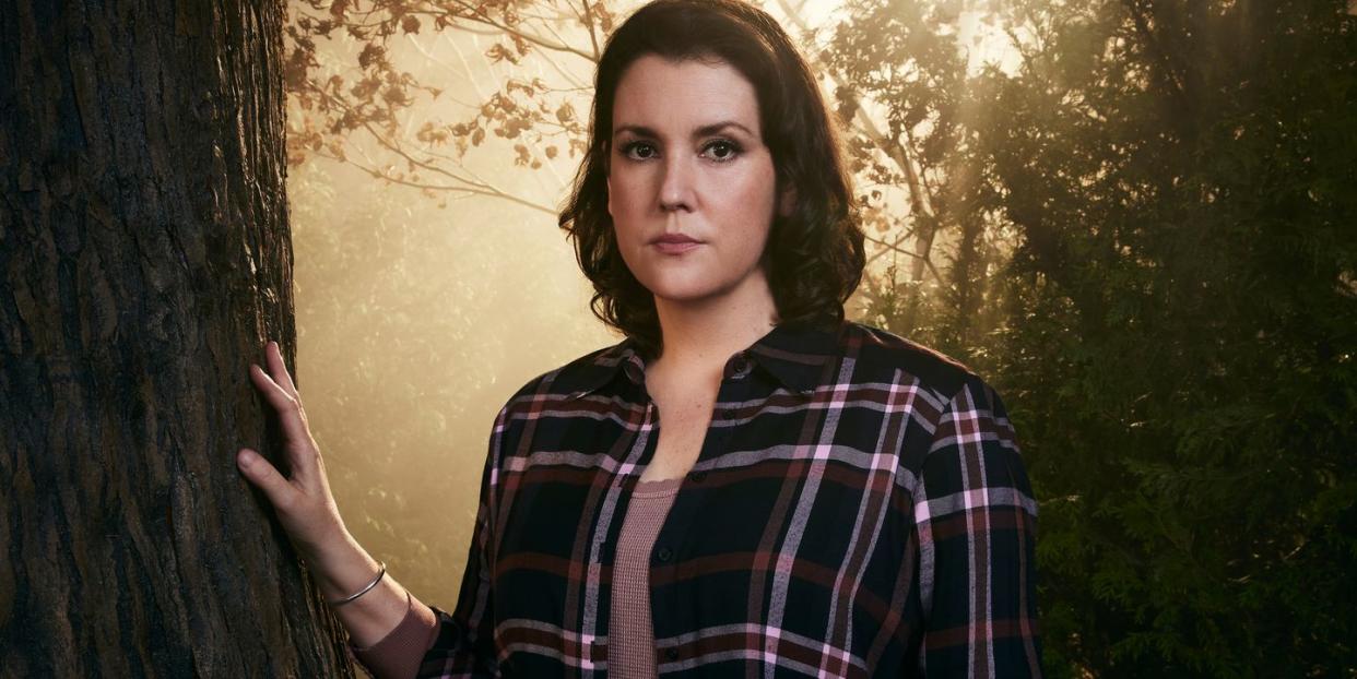 yellowjackets star melanie lynskey, in character as shauna, stands in a forest with her right hand resting on a tree trunk