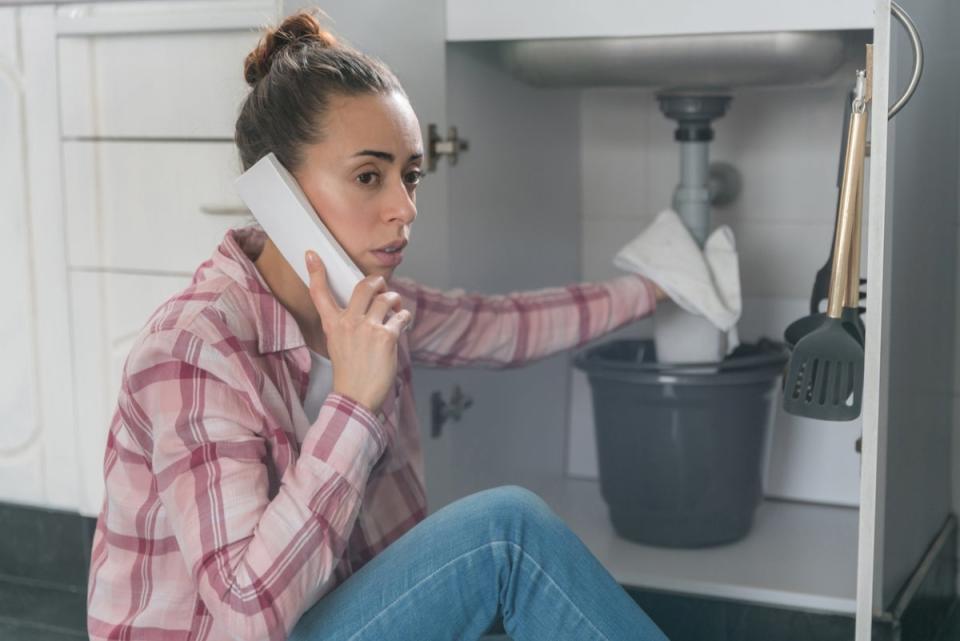 Woman holding a cordless telephone to her ear, holding a towel to plumbing under the sink.