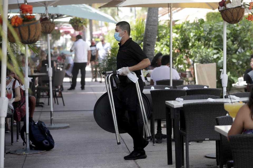 A waiter wears a protective face mask and gloves while working at the il bolognese restaurant along Ocean Drive during the coronavirus pandemic, Sunday, July 12, 2020, in Miami Beach, Fla. Restaurants throughout Miami-Dade County are open only for outdoor dining, take-out and delivery. (AP Photo/Lynne Sladky)