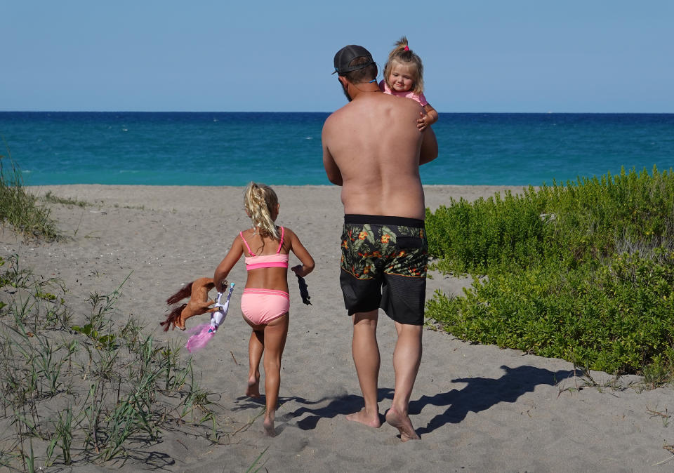 JENSEN BEACH, FLORIDA - MAY 04: Cole Hunter carries Harper Hunter, 1, as Holly Hunter,4, follows along as they arrive at the beach on May 04, 2020 in Jensen Beach, Florida. Restaurants, retailers, as well as beaches and some state parks reopened today with caveats, as the state continues to ease restrictions put in place to contain COVID-19. The counties of Palm Beach, Broward and Miami Dade continue to maintain restrictions. (Photo by Joe Raedle/Getty Images)