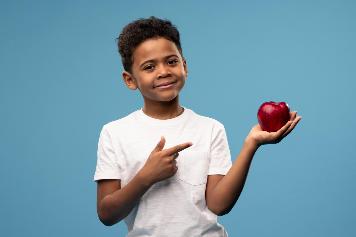 Happy little African boy with big red ripe apple in hand pointing at the fruit and looking at you while recommending it as healthy food