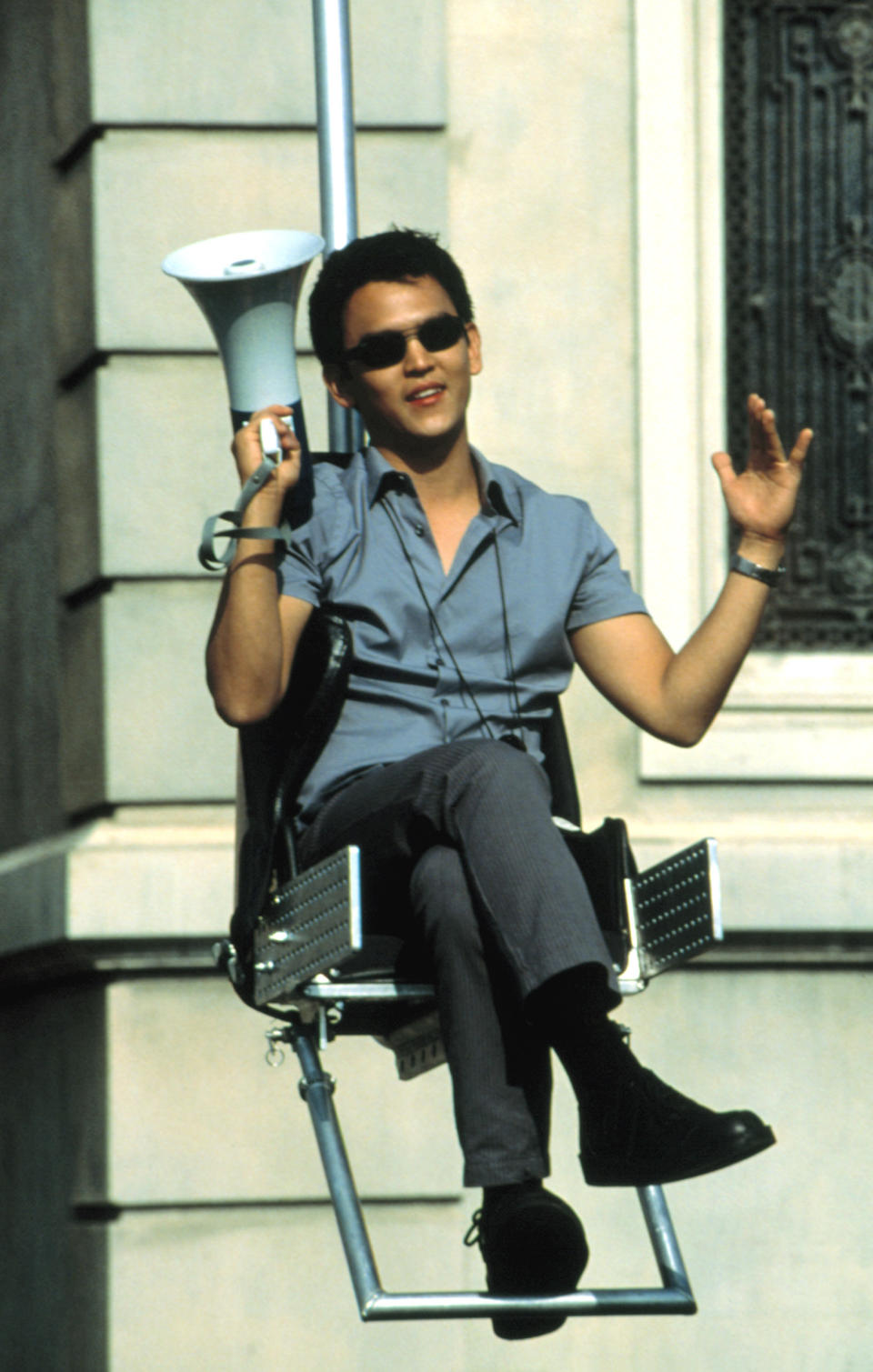 John Cho, sitting in a chair lifted by a metal structure, holding a megaphone and gesturing with his other hand, wearing casual attire and sunglasses