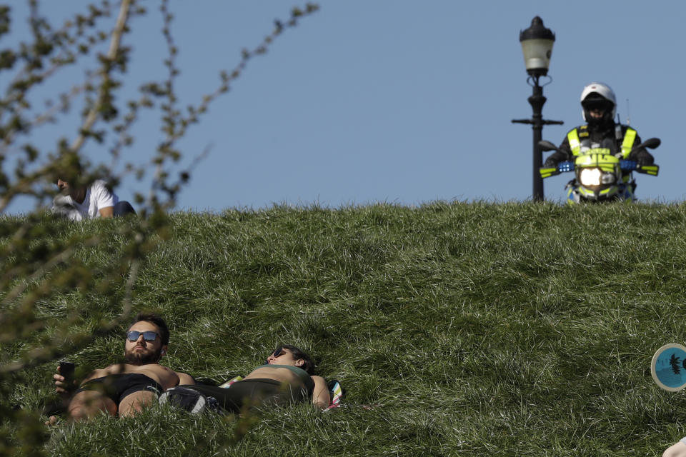 A man and woman lay down sunbathing as a police officer on a motorbike shouts for people who were relaxing in the sun to go home, on Primrose Hill in London, to stop the spread of coronavirus and keep the park open for people observing the British government's guidance of social distancing, only using parks for dog walking, one form of exercise a day, like a run, walk, or cycle alone or with members of the same household, Sunday, April 5, 2020. The new coronavirus causes mild or moderate symptoms for most people, but for some, especially older adults and people with existing health problems, it can cause more severe illness or death. (AP Photo/Matt Dunham)