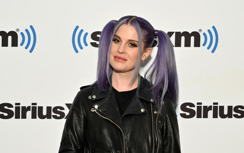 Kelly Osbourne wearing a black leather jacket, with purple hair, standing before a SiriusXM backdrop