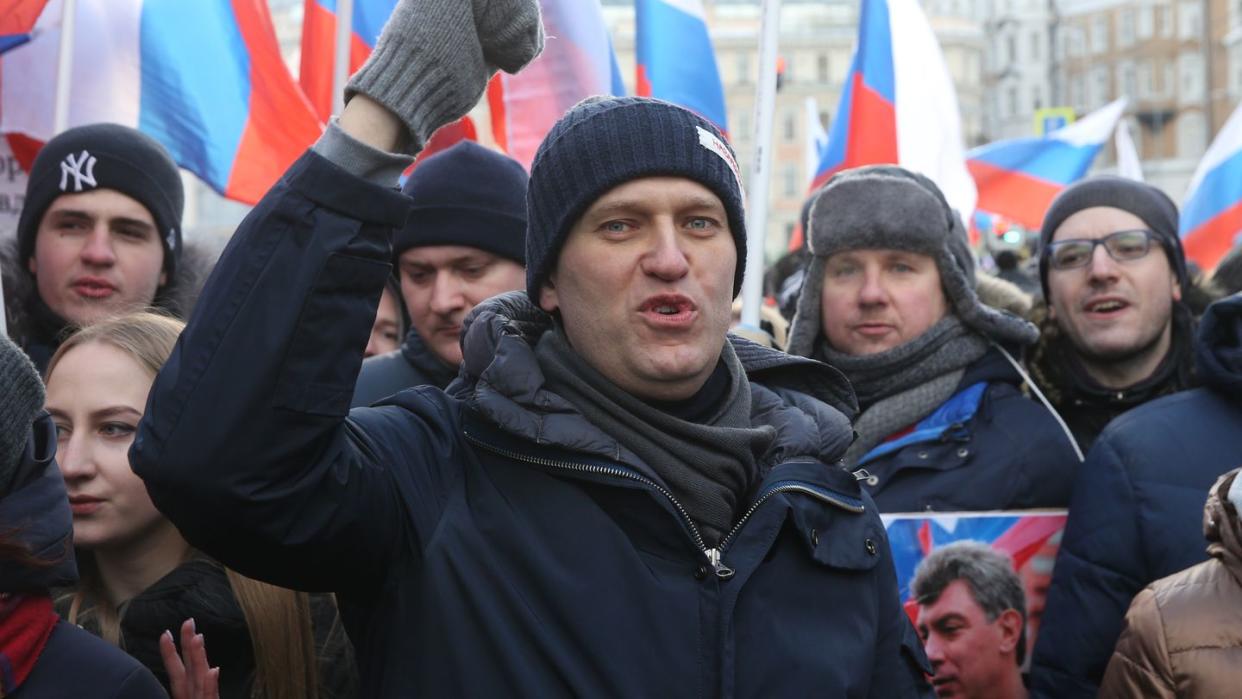 alexei navalny chants and holds one hand above his head as he stands in front of a group of people, he wears a hat, mittens, scarf and coat, several russian flags are behind