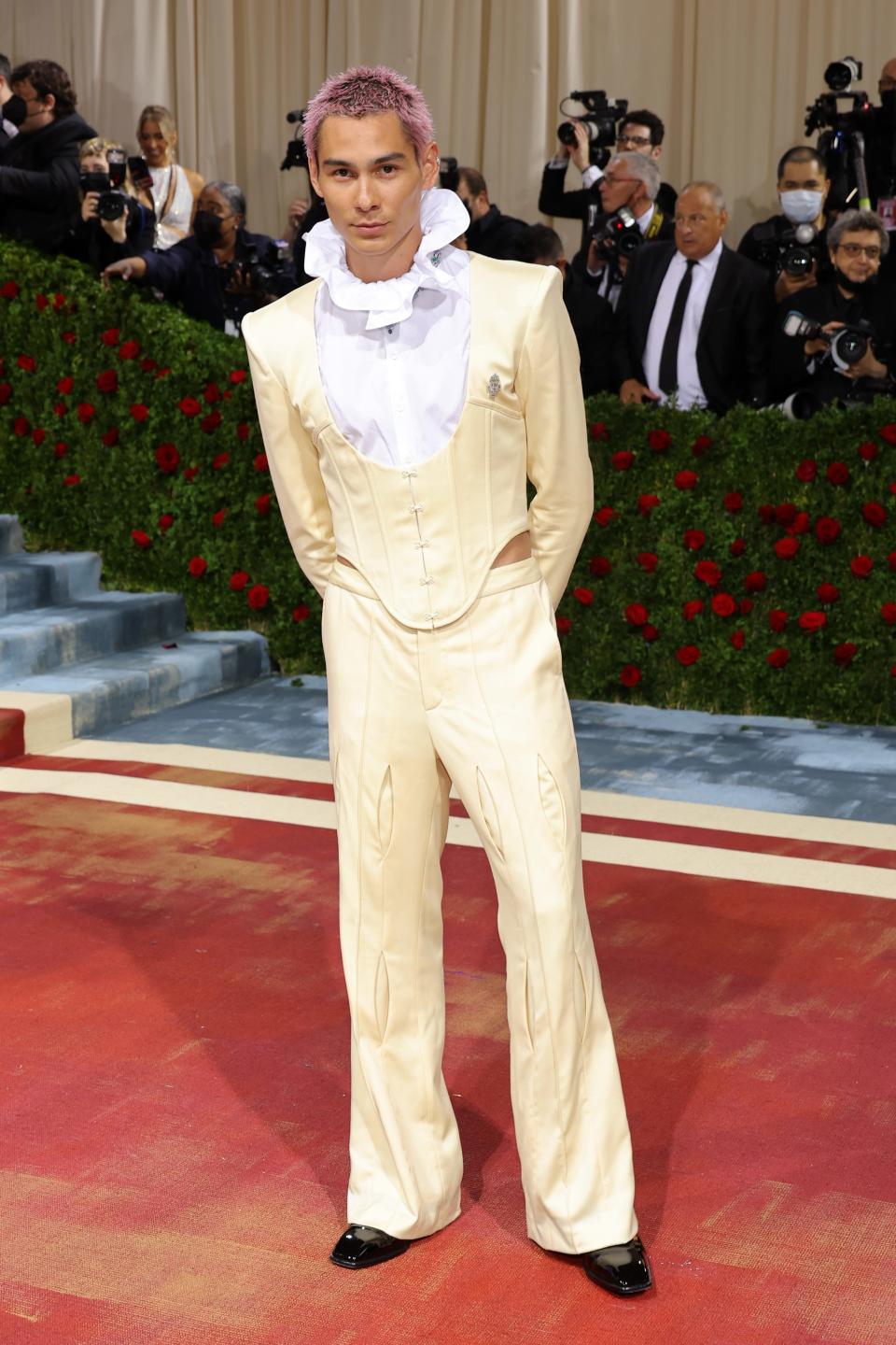 Evan with pink dyed spikey hair and a beige cream suit that has a corset-like vest jacket and a white button down underneath with a large ruffled collar.