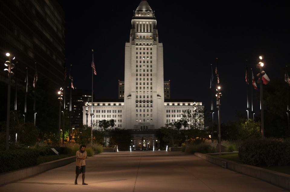 A barefooted homeless person wanders through Gloria Molina Park, with the illuminated Los Angeles City Hall looming in the background in Los Angeles, Tuesday, Sept. 12, 2023. Billions of dollars have been spent on homelessness in the region, and an array of new programs are in place. But Mayor Karen Bass says it's possible that the number of homeless people will continue to increase, in part because of evictions and the end of pandemic aid that helped pay bills for lower-income households. (AP Photo/Jae C. Hong)