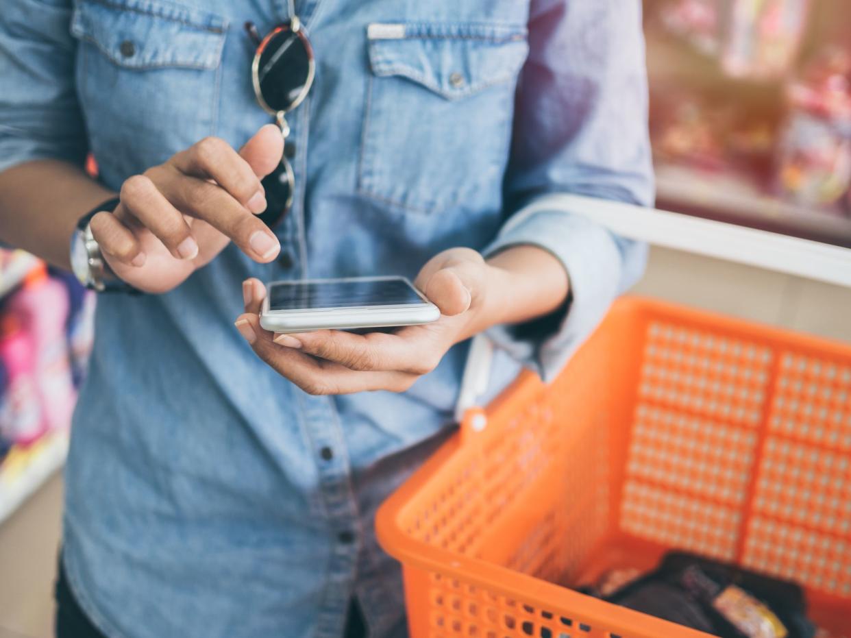 Woman wearing blue jeans shirt and sunglasses using mobile phone to compare price and holding orange shopping basket in mini mart background.