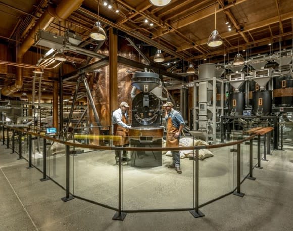 Two people at a coffee roaster in a building with exposed pipes in the ceiling.