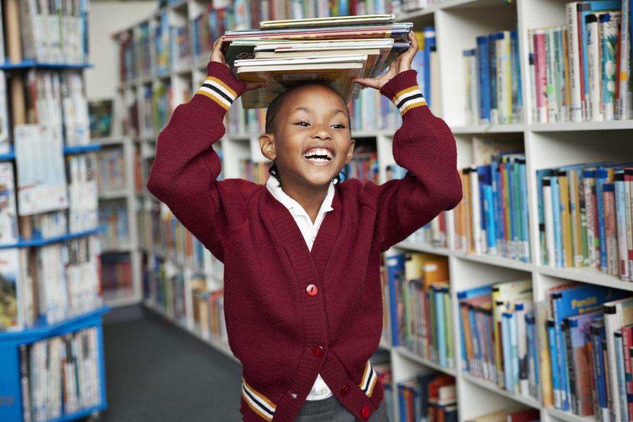 cute schoolgirl smiling balancing stack of books on the head at library