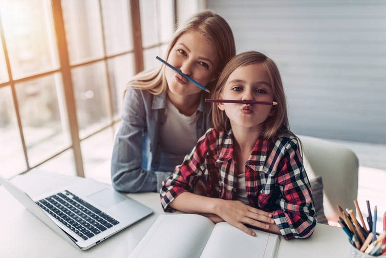 A woman and child sit at a desk doing homework, holding pencils like mustaches under their noses