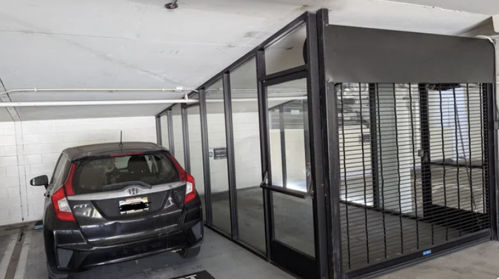 A small black car is parked in a parking space next to a locked glass and metal storage cage in an underground parking garage