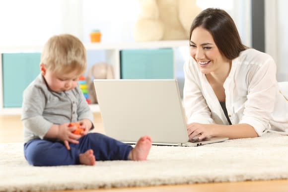 Baby playing with a red toy while his mother smiles at her laptop's screen.