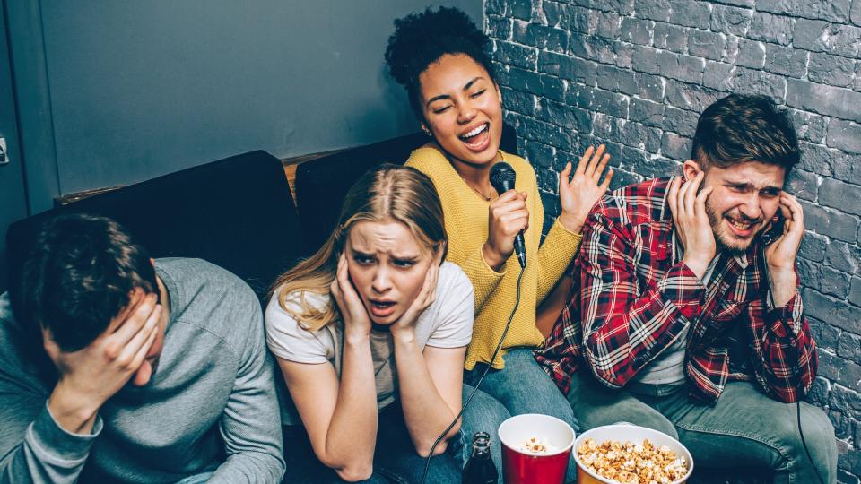 Young woman singing badly whilst her three other friends cover their ears.