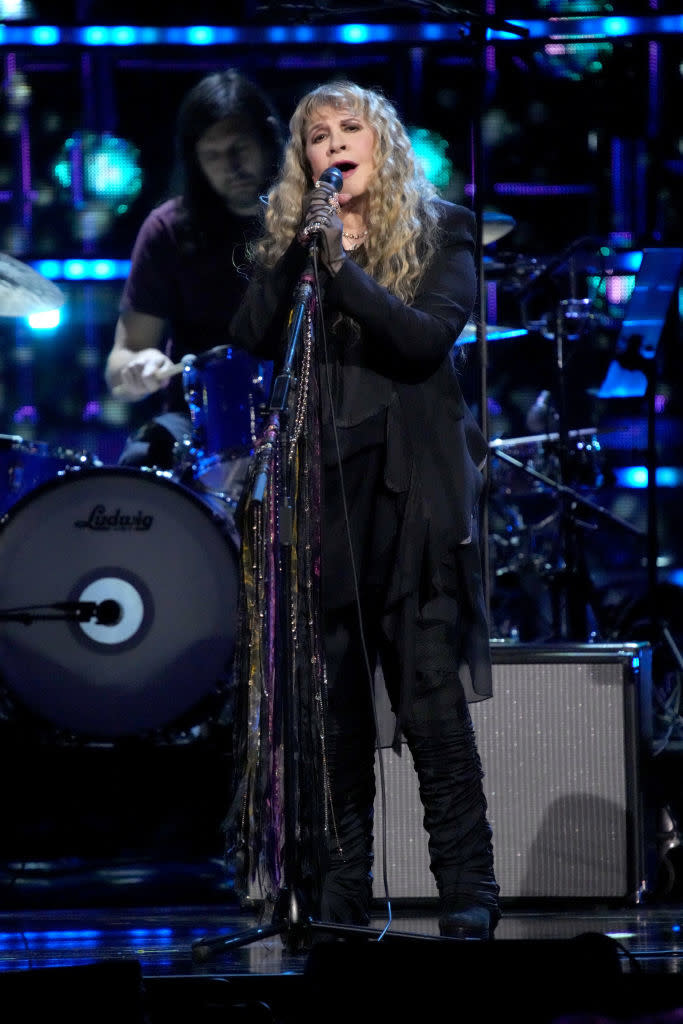 Stevie performing on stage with microphone, wearing layered clothing and boots, with band members in background