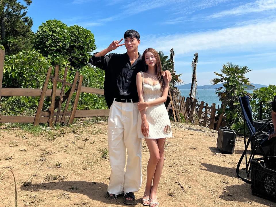 a man makes a v pose next to a woman holding her arm on a sandy tree-lined beach