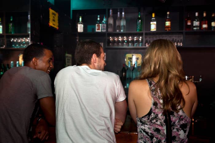 Three people are sitting at a bar, viewed from behind. The bar shelves are stocked with various bottles of liquor and glasses