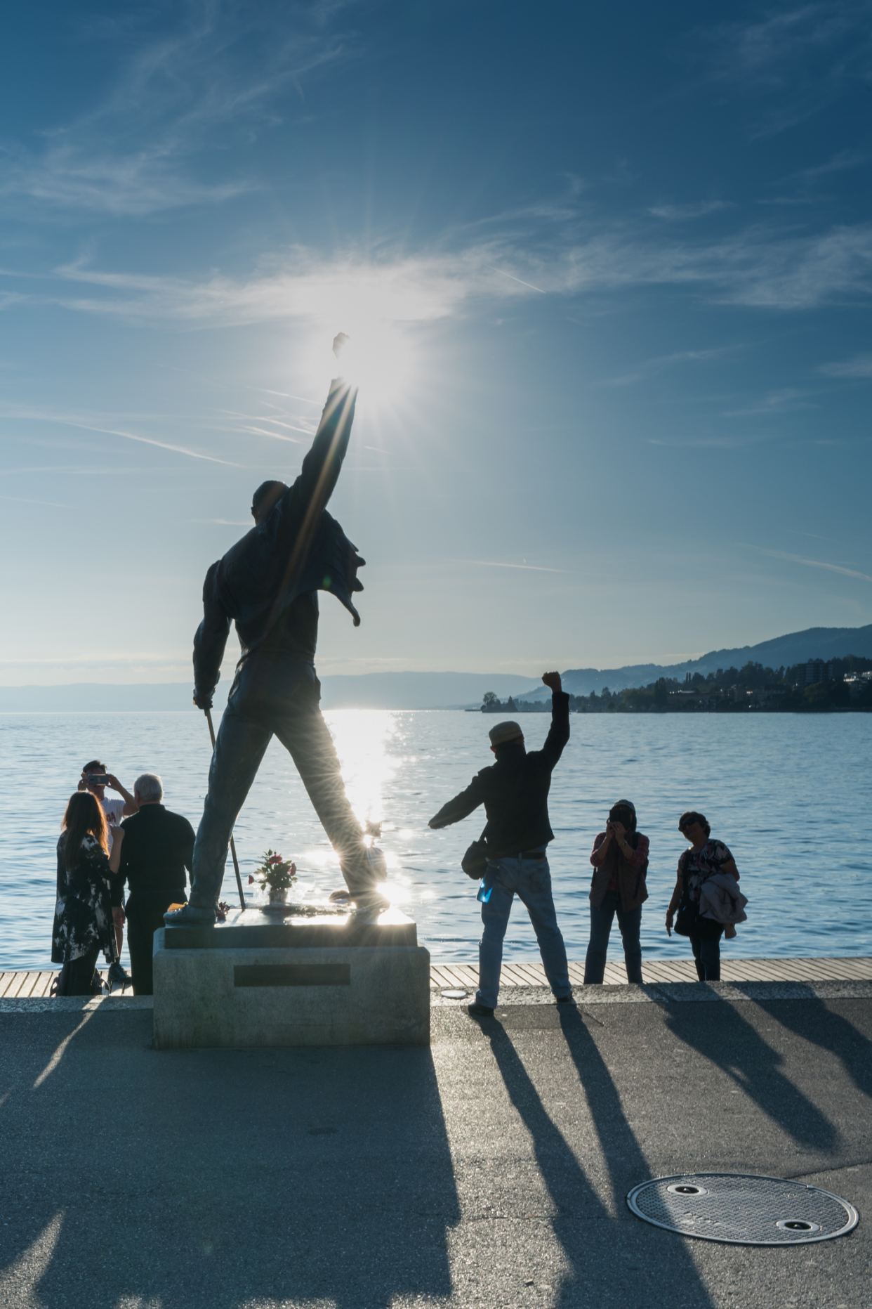 Freddie Mercury's Memorial Statue, Montreux, Switzerland overlooking Lake Geneva with the sun's light dramatically touching the tip of his fist with a few people on the edge, one man is copying Mercury's stance