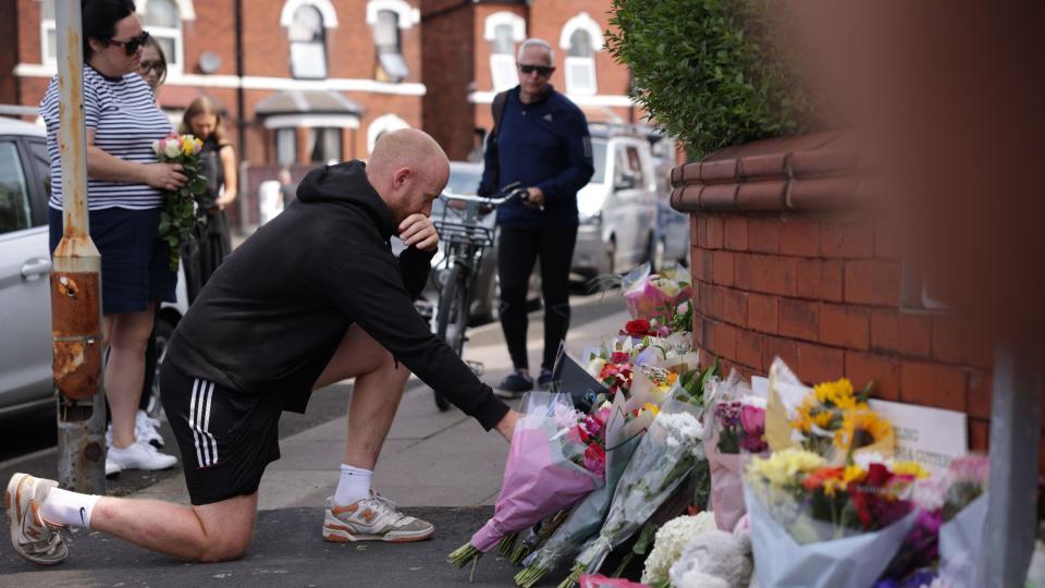 A man kneels down to read tributes left at the scene. An array of flowers can be seen and more people stand in the background.