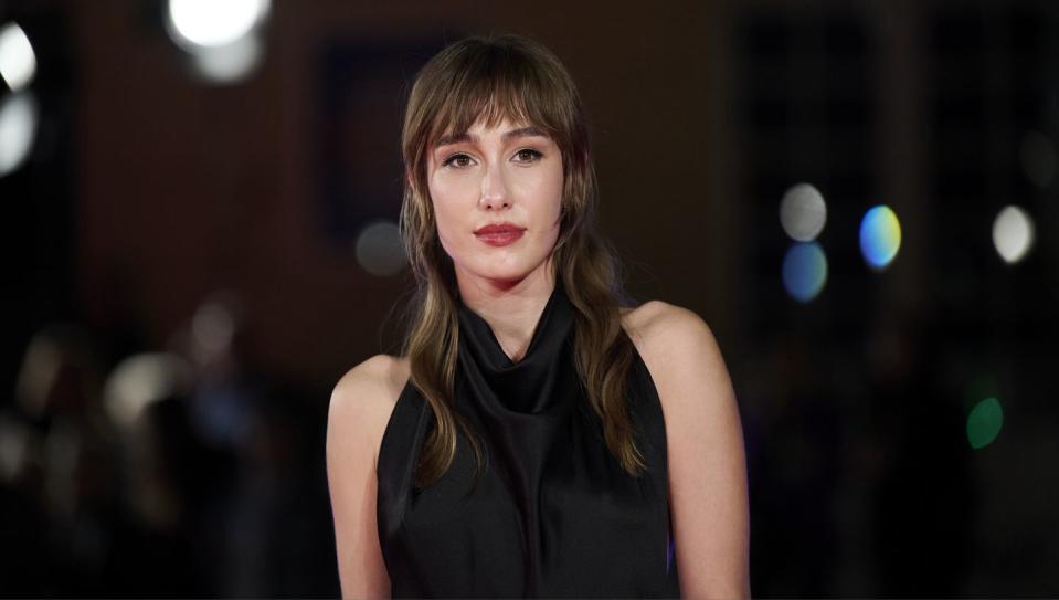 almudena amor, a woman stands looking at the camera with a neutral facial expression, she has brown hair worn in loose waves with a fringe, she wears a black halterneck dress