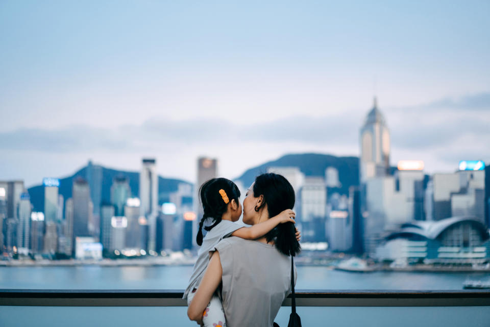 A woman holds a young girl on her shoulder, both facing a city skyline with tall buildings and mountains in the background