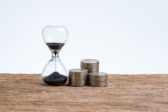 An hourglass sitting on a table next to three stacks of coins.