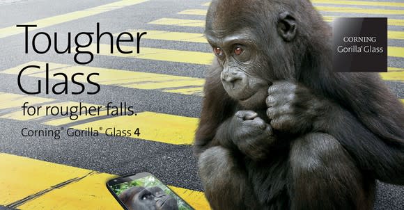 A young gorilla sitting on a city street next to a smartphone with the caption "Tougher Glass for rougher falls"