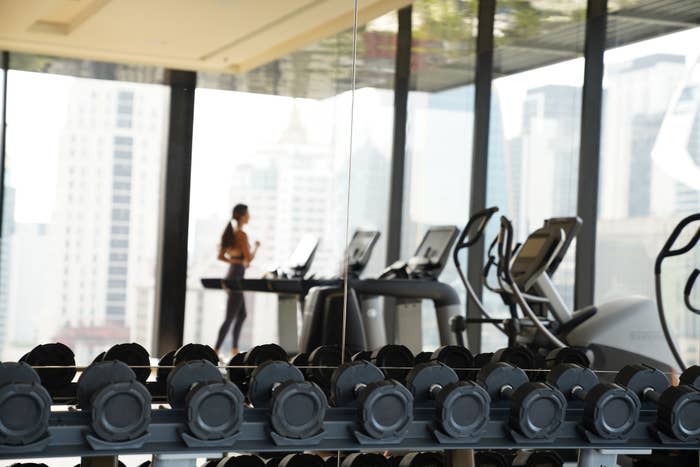 Dumbbell rack in a modern gym with a person running on a treadmill in the background, city buildings visible through large windows