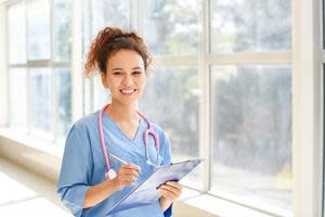 Photo purchased by Workwolf. Description for visually impaired: A smiling woman in blue scrubs faces the camera at a diagonal, holding a clipboard and pen. She is standing in a sunny hallway, and has a stethoscope around her neck and a curly ponytail hairdo.