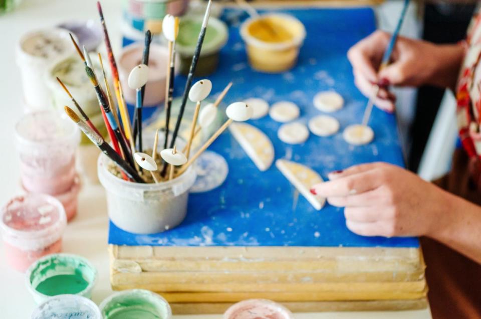 A woman paints small ceramic objects at her painting station setup with different paints and paint brushes. 