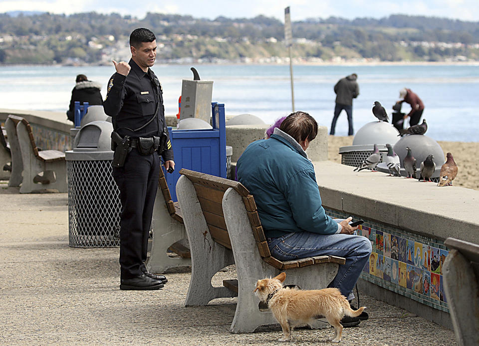 Police Sgt. Leo Moreno tells a couple they have to keep moving and can't sit on the Esplanade bench Wednesday, March 25, 2020, in Capitola, Calif., explaining that the bench may have the coronavirus on it. (Dan Coyro/The Santa Cruz Sentinel via AP)