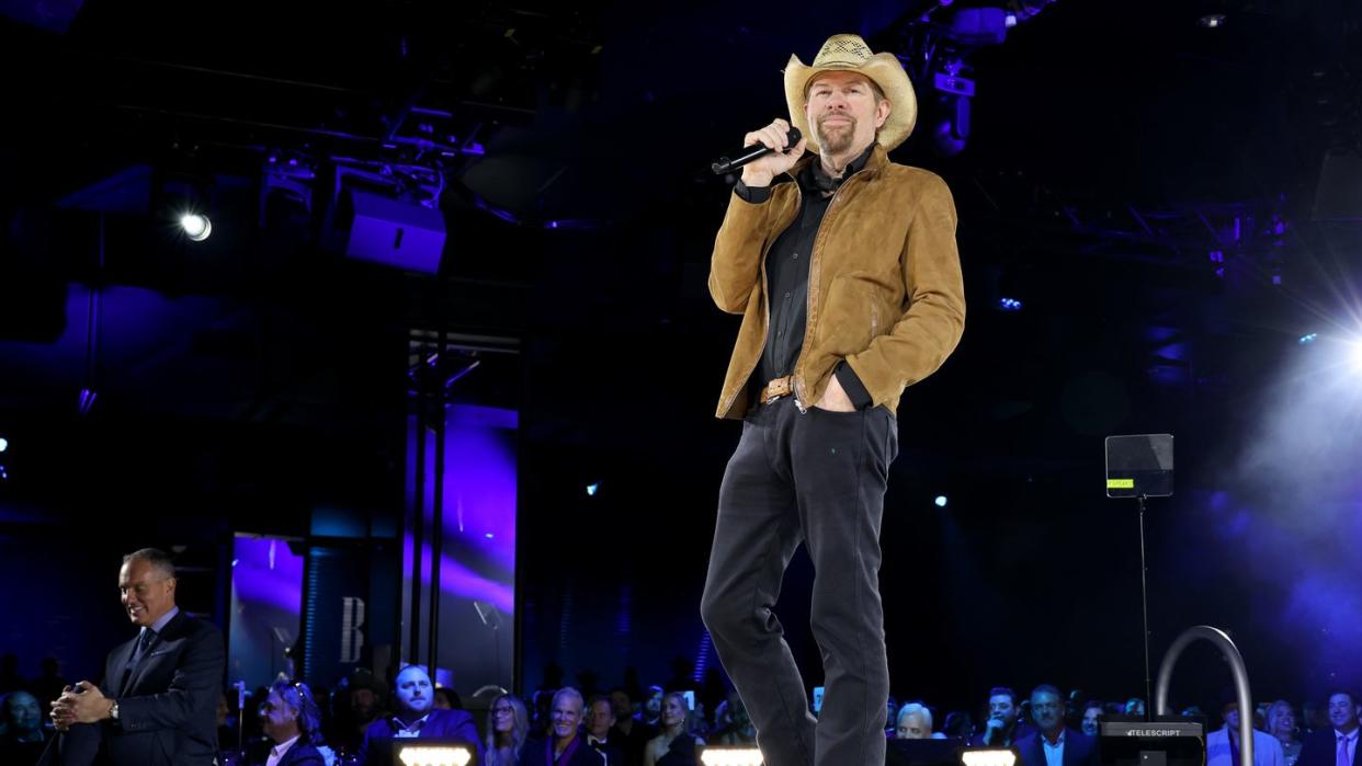 toby keith smiles as he holds a wireless microphone near his face, he wears a cowboy hat, brown jacket, black shirt and black pants