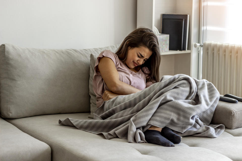 Woman holding hands on stomach suffering from stomach pain with eyes closed. She sits on a couch with a blanket over her knees.