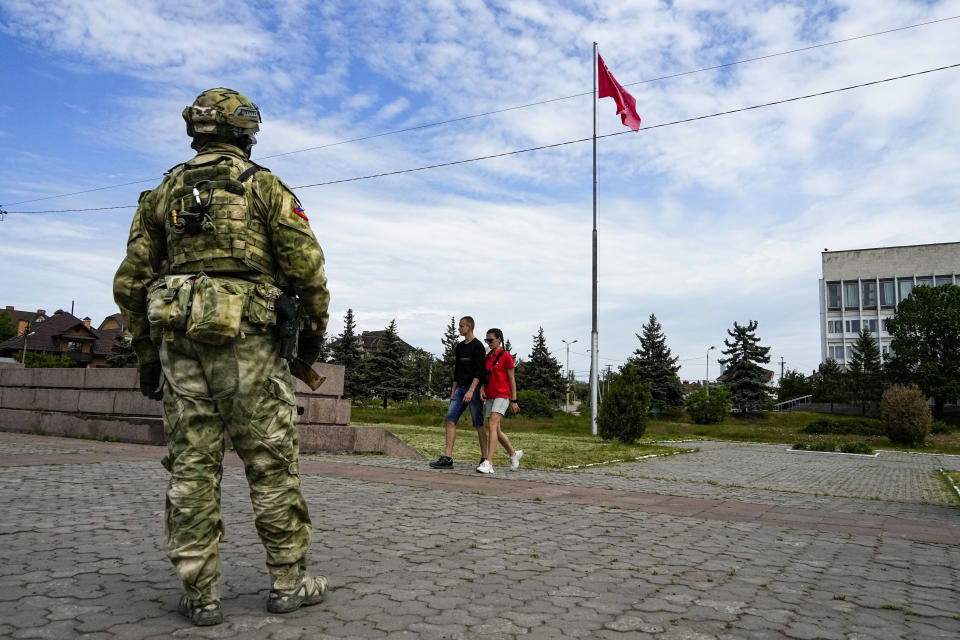 FILE - A young couple walks past a Russian soldier guarding an area at the Alley of Glory exploits of the heroes - natives of the Kherson region, who took part in the liberation of the region from the Nazi invaders, in Kherson, Kherson region, south Ukraine, Friday, May 20, 2022, with a replica of the Victory banner marking the 77th anniversary of the end of World War II right in the background. The Kherson region has been under control of the Russian forces since the early days of the Russian military action in Ukraine. This photo was taken during a trip organized by the Russian Ministry of Defense. (AP Photo, File)